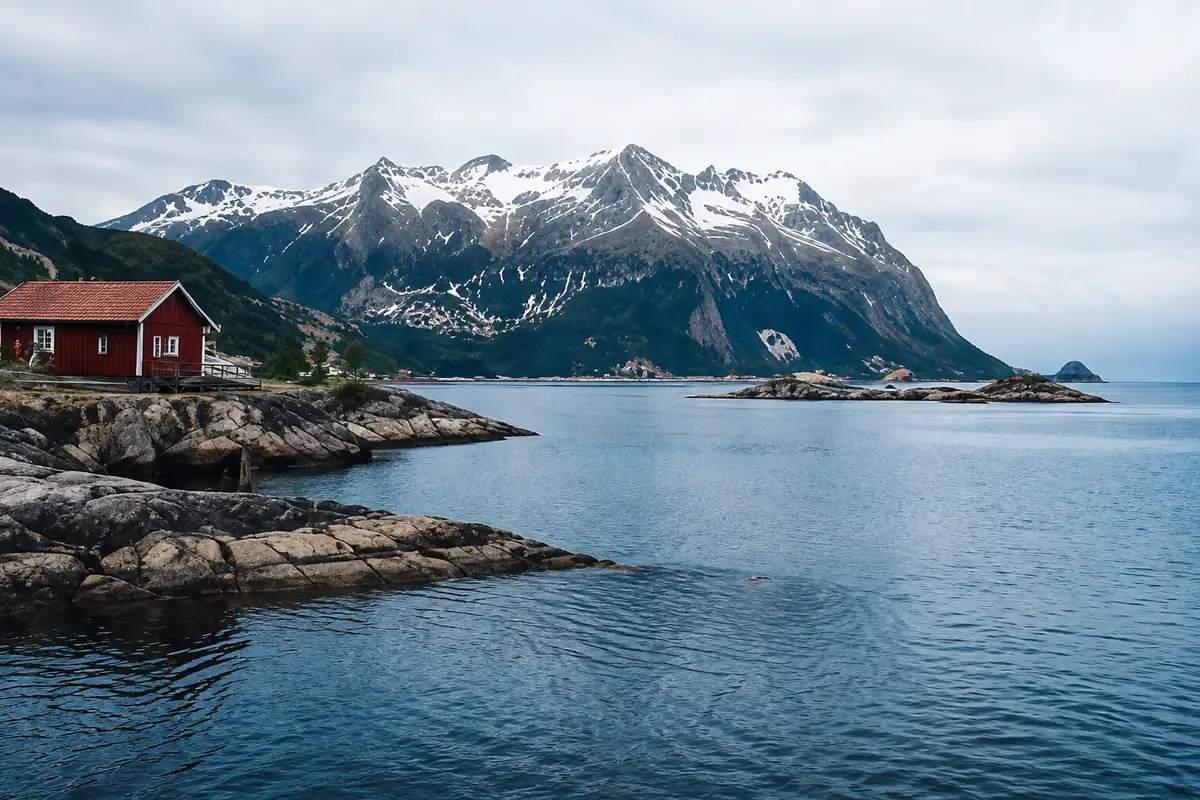 Scenic Norwegian coastal landscape with snow-capped mountains, rocky shoreline, and a traditional red fishing house.